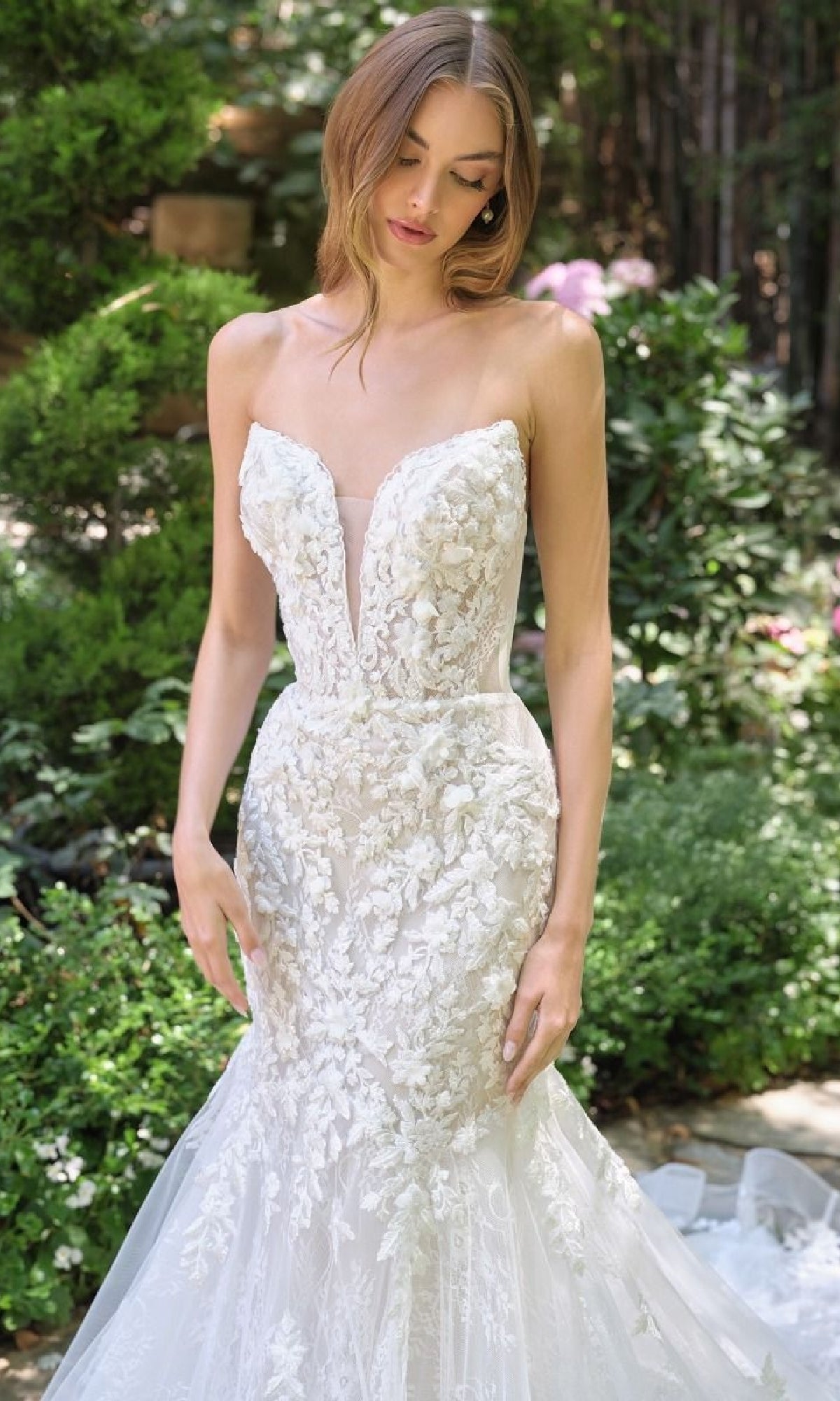 Wearing the Strapless Mermaid Bridal Dress: Andrea & Leo WL088, a woman stands outdoors among greenery, looking down.